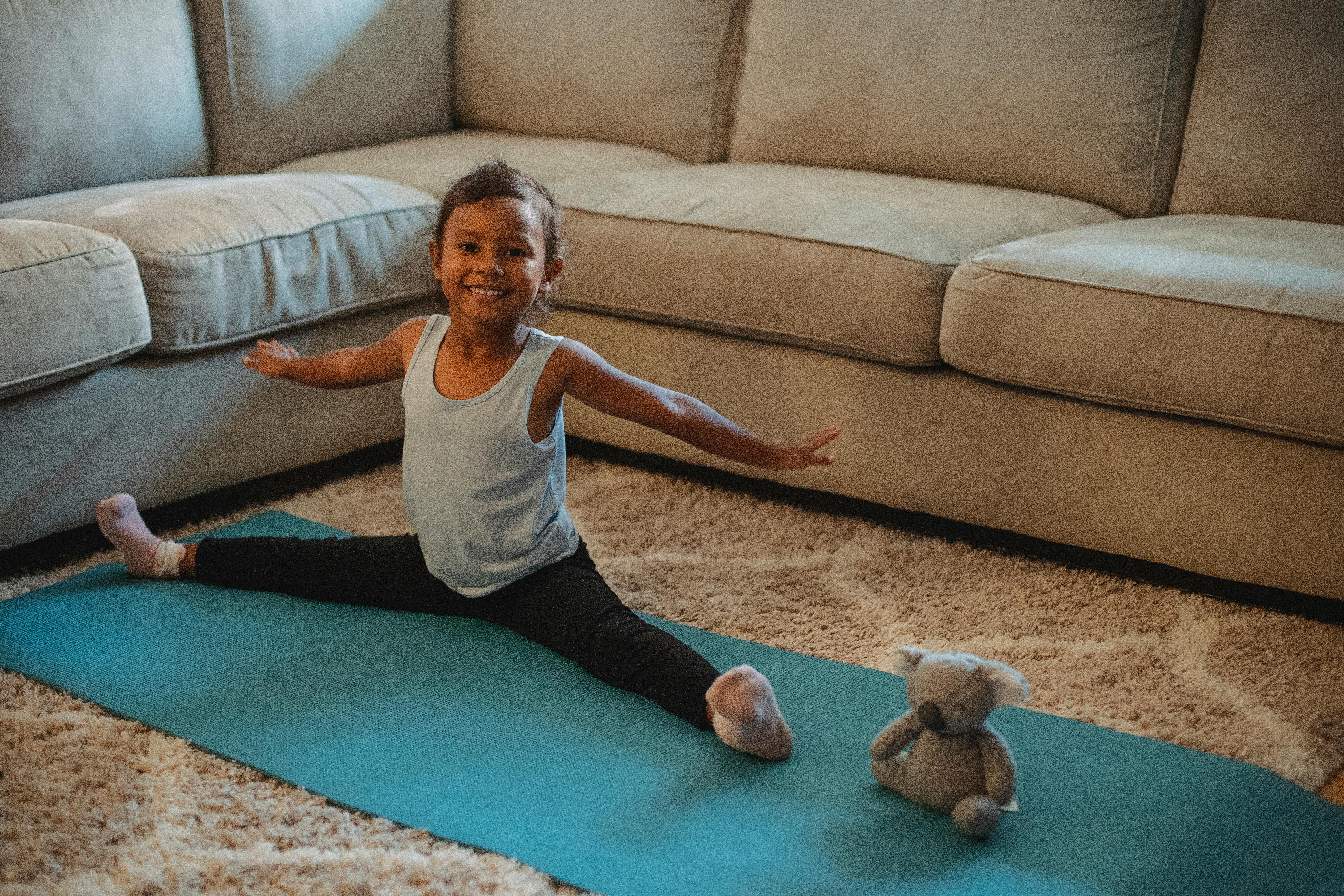 Cheerful little ethnic little girl stretching on stretch mat at home with toy and looking at camera