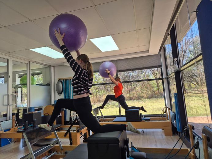 Two women lunging on reformers holding exercise balls overhead
