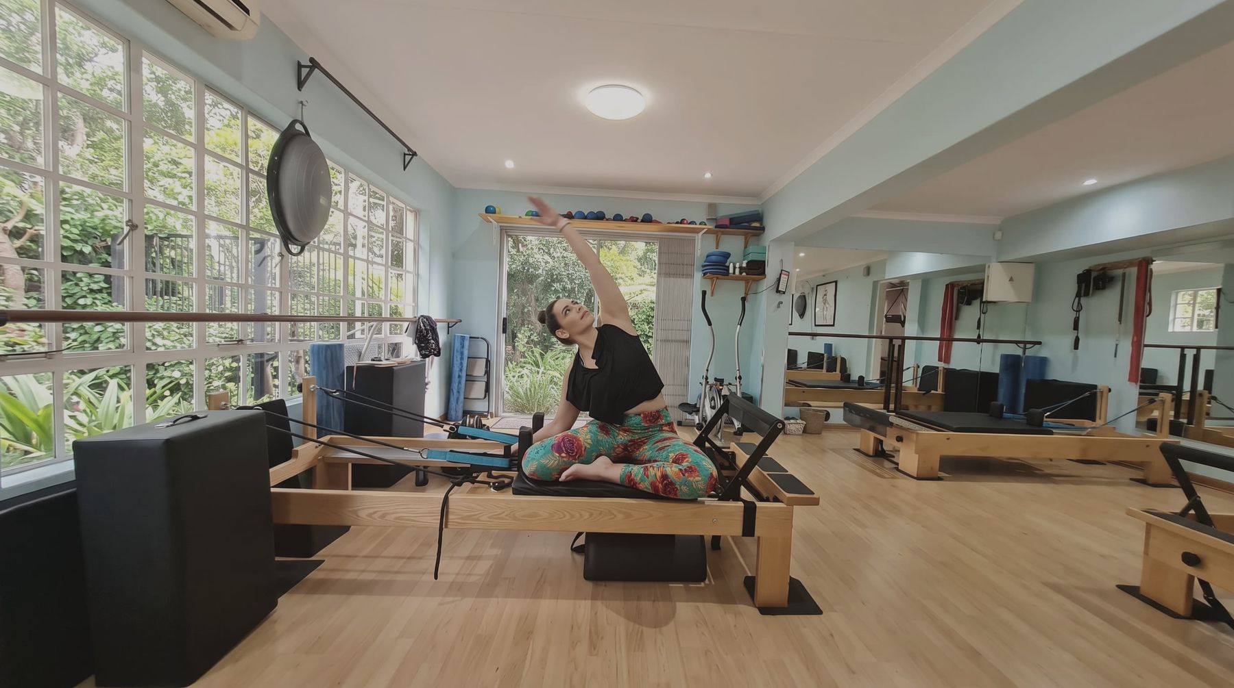 An instructor on the reformer in a side stretch, with the garden through the studio windows behind her