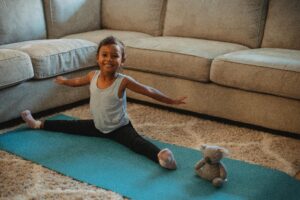 Cheerful little ethnic little girl stretching on stretch mat at home with toy and looking at camera